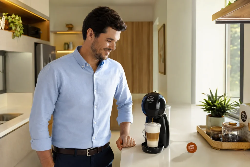 Um jovem profissional de camisa social sorrindo na cozinha de seu apartamento enquanto observa sua Dolce Gusto preparar um Cappuccino em uma caneca de vidro. Cafeteira Dolce Gusto vale a pena